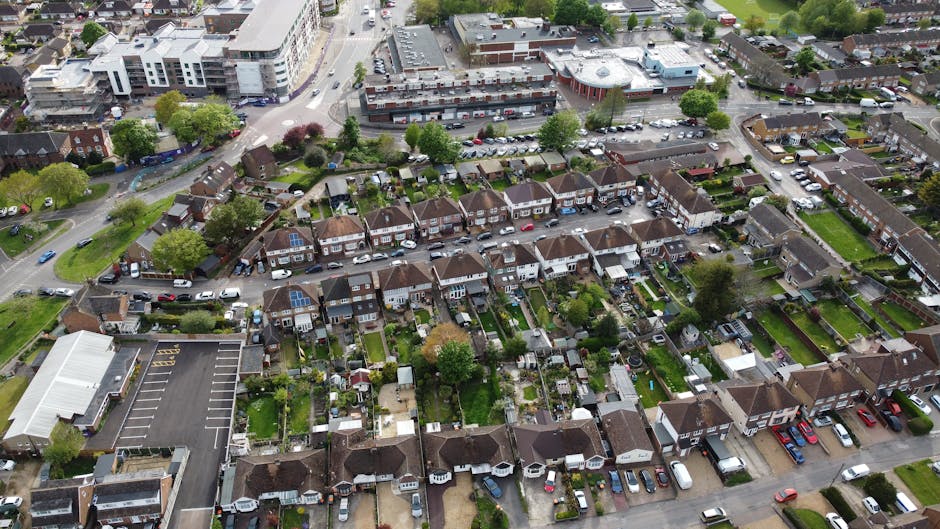 An aerial view of a residential neighbourhood in Golders Green, showing a row of terraced houses with small private gardens at the back, many with green lawns, trees, and garden sheds. Narrow paved pathways separate the gardens from the houses, and several cars are parked along the driveways and streets. In the foreground, a larger commercial or community building with a white roof is visible. Surrounding the residential area are roads with more parked cars, and beyond that, larger buildings and parking lots, indicating urban infrastructure. The scene depicts a typical suburban setting suitable for house removals and furniture transport, with clear visual evidence of the logistical activities involved in home relocation and packing and moving processes. Man with Van Golders Green occasionally supports these services in the local area.