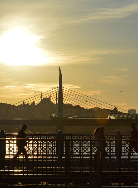 A sunset scene over a river showing the Millennium Bridge in central London with its tall, curved central pylon and suspension cables. In the foreground, a black metal pedestrian railing runs along the riverside walkway, with four silhouetted individuals walking and standing near the railing, some engaged in conversation and others looking out over the water. Behind the bridge, the cityscape includes low-rise buildings with rooftops and chimneys, and the sky displays a warm, golden hue with wispy clouds illuminated by the setting sun. The overall scene captures a peaceful moment during early evening, highlighting London’s iconic architecture and riverside environment, relevant to home relocation and moving logistics discussions.