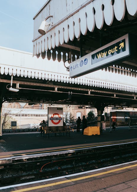 A person in casual clothing is seen inside a train station, standing on a platform next to a red passenger train that is parked alongside the tracks. The platform has a weathered, blue-painted metal c