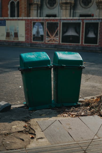 Two green wheelie bins are positioned side by side on a pavement next to a curb, with one slightly behind the other. They are made of plastic and have black handles. The bins are situated in an urban environment, with a partially visible brick and glass building facade across the road and a large window in the background. The pavement includes a section of concrete tiles, and there is some loose debris and dried leaves near the base of the bins. This scene captures a typical city street setting, relevant to house removals and waste disposal services offered by Man with Van Golders Green, with the bins possibly representing waste management options during a home relocation or moving process.