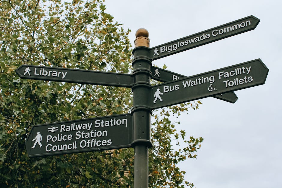 A black multi-directional street signpost situated outdoors against a backdrop of green-leaved trees and a cloudy sky. The signpost features six directional signs, each with white text and symbols, indicating nearby locations such as Biggleswade Common, Library, Bus Waiting Facility, Toilets, Railway Station, Police Station, and Council Offices. The signs are mounted on a pole with a rounded top cap. In the context of house removals and home relocation services, this image illustrates navigation and directional guidance relevant to the Golders Green area, where Man with Van Golders Green provides professional removals and moving logistics including efficient furniture transport and packing support.