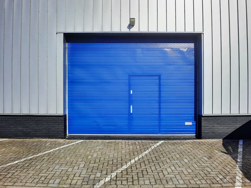 A large blue industrial roller shutter door is situated on the exterior of a commercial building with vertical grey metal paneling. Above the door, a small security light is mounted on the wall. The ground is paved with concrete bricks, with white line markings indicating designated parking spaces directly in front of the loading bay. The area appears tidy and well-maintained, suitable for furniture transport and home relocation activities, as carried out by Man with Van Golders Green. The scene provides a clear view of a typical loading area used in house removals, where trucks or vans can easily access the interior for loading or unloading household items. The absence of visible furniture or packing materials suggests the image depicts an entry point prepared for a moving process, with the focus on the loading bay´s structural features and environment conducive to efficient relocations.