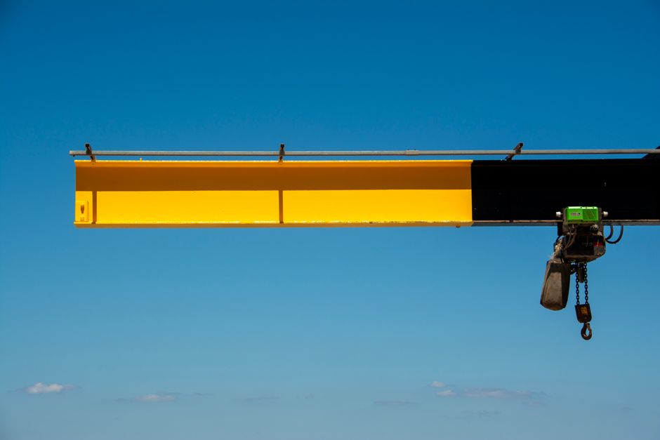 A close-up view of a yellow and black crane jib arm extending horizontally against a bright blue sky with a few small, scattered clouds. The yellow section is prominent, with the black part partially visible at one end. Attached to the crane is a chain hoist with a black casing, hanging down on the right side, connected to the crane's trolley mechanism. The scene is outdoors, likely at a construction or loading site near a property, and the image captures the structural elements used for heavy lifting in moving or construction activities, relevant to home relocation or furniture transportation services. The image emphasizes the equipment used during loading or unloading processes, possibly at a house removal site located in NW11 or Golders Green, aligning with the themes of removals and moving logistics provided by Man with Van Golders Green.