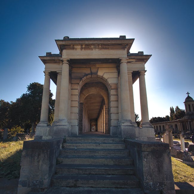 A stone entrance with a set of concrete stairs leading up to a large archway supported by four columns, situated outdoors on a sunny day. The structure is part of a historic or memorial building, with trees and other cemetery features visible to the sides. The stairs and platform are weathered, showing signs of age. Behind the structure, there is a pathway lined with a series of columns or pillars, suggesting a walkway through a memorial site. Natural lighting highlights the stone textures and architectural details, indicating a daytime setting. This scene is related to house removals or transportation logistics, fitting with the context of moving or packing items for relocation, potentially near Golders Green Crematorium, as per the page title. Man with Van Golders Green often facilitates such home relocation and furniture transport services, including loading and protective packing processes that might involve stairs and stonework similar to this structure.