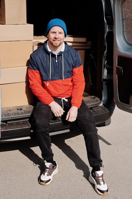 A man wearing a white shirt and black cap is in the process of loading brown cardboard boxes into the back of a large grey cargo van, which is parked outdoors on a paved surface. The van's rear doors 
