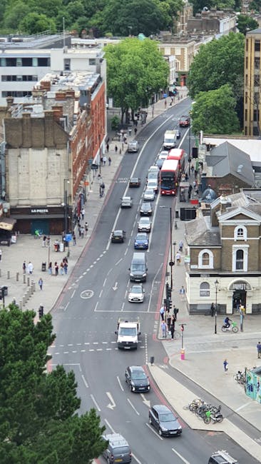 An aerial view of a busy urban street in Golders Green, showing multiple lanes with cars and vans moving in both directions. On the left side, there are commercial buildings with shopfronts and pedestrians walking on the pavement. On the right side, there is a small, standalone building with a classical architectural style and a bicycle parked nearby. The street is bordered by several large green trees, providing shade along the sidewalk. Visible in the distance are residential and commercial structures, and the overall scene depicts typical city traffic during daytime. This environment illustrates the context in which Man with Van Golders Green facilitates home relocations and furniture transport, involving loading and unloading processes within a bustling urban setting.